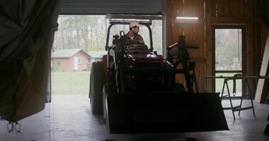 Young man of European appearance with a beard, starts a small agricultural tractor with a bucket and drives it out of the garage, backing it up, going to work on the farm on an autumn day photo