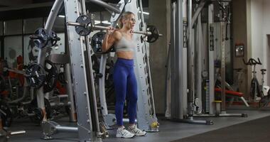 Young woman of athletic physique, dressed in a sports uniform, squats with a barbell on the exercise machine, doing in the gym. filming from the side, general plan of the gymnasium photo
