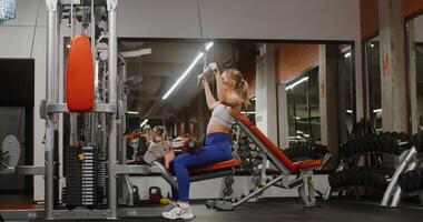 Blonde woman, dressed in sports uniform, lifts weights while exercising on the exercise machine in the gym alone, the exercise machine is fully visible photo
