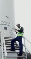 A power plant worker in a green vest and hard hat uses a tablet with a serious look while standing on the steps near the entrance to industrial wind generator, vertical . in 4k, red komodo photo