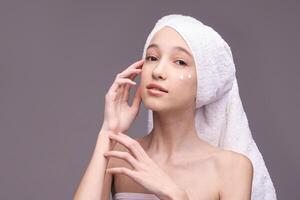 Young caucasian female applying skincare cream with towel on head in minimalist setting. photo