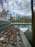 Old Boats Resting on the Shore Near a Calm Pond Surrounded by Trees Under a Cloudy Sky in Early Spring photo