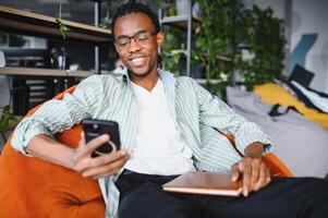Smiling university student using smartphone and holding book in modern library photo