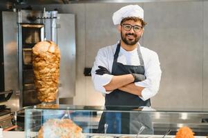 Chef smiling with arms crossed in front of shawarma rotisserie and display case photo