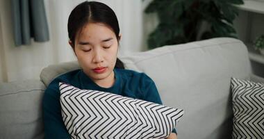 Handheld high angle view shot, Young Asian woman is sitting on a sofa hugging a pillow, lost in thought, pondering life problems in a quiet room at home. Mental health, anxiety, depression video