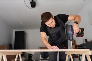 Man in work overalls bib using cordless power drill to make hole in laminated wood board. Male carpenter working in home workshop photo