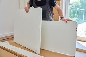 Man in work overalls assembles white furniture panel near unpacked parts and tools at home. Handyman preparing part of bed for assembly in bedroom photo