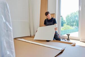 Man in work overalls assembles white furniture panel near unpacked parts and tools at home. photo