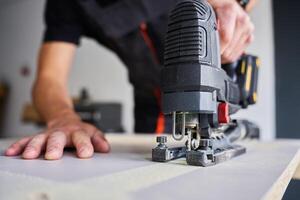 Worker in workwear using handheld cordless electric jigsaw to cut wooden board in garage. Skilled carpenter working in workshop photo