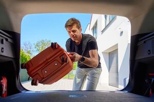 Man placing orange rolling suitcase into open vehicle trunk, preparing for journey or vacation on sunny day. Inside view of traveler loading luggage in car. Travel or weekend road trip concept photo