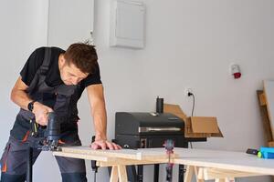 Carpenter cuts wooden board with electric jigsaw in home workshop. Man in workwear working with wood on workbench in garage. Woodworking and carpentry concept photo