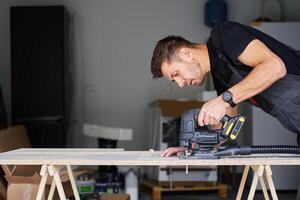 Person in workwear using handheld cordless electric jigsaw to cut wooden board in garage. Skilled carpenter working in workshop photo