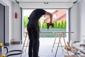 Worker using power drill on wooden board supported by sawhorses in home garage workshop. Man working in workshop, drilling hole in chipboard for furniture assembly photo