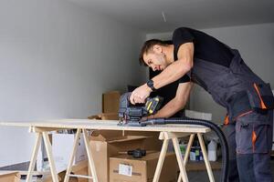 Worker in workwear using handheld cordless jigsaw to cut wooden board in garage. Skilled carpenter working in workshop. Man doing DIY home renovation photo