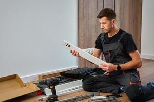 Man in overalls assembles furniture, examining white panel and surrounded by tools. Handyman in work clothes preparing parts pf cabinet to assembly. DIY furniture project photo