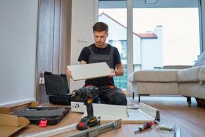 Man assembles furniture, examining white panel and surrounded by tools. Handyman in work clothes preparing parts pf cabinet to assembly. DIY furniture project photo