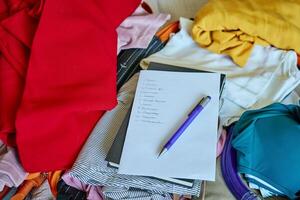 Handwritten checklist and pen on top of colorful folded clothes inside open suitcase. Concept of travel planning and packing essentials for trip photo
