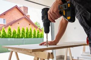 Man in using cordless power drill to make hole in laminated wood board. Male carpenter working in home workshop, drilling chipboard on workbench photo