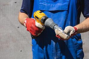 A worker in a blue jumpsuit holds an electric screwdriver with a riveter attachment and an exhaust rivet against a slate wall photo