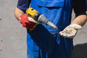 A man in a blue jumpsuit holds electric screwdriver with a riveter attachment and exhaust construction rivets on the background of a slate wall photo