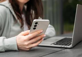 A person using a smartphone while sitting with a laptop on a wooden table outdoors, symbolizing multitasking and modern technology. photo
