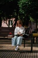 Writing ideas, into ,notepad. Schoolgirl is sitting on the bench with a bunch of books outdoors photo