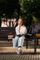 Writing, ideas ,,into notepad. Schoolgirl is sitting on the bench with a bunch of books outdoors photo