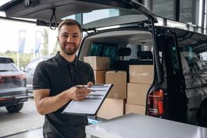 Pen, and ,the, notepad in hands. Delivery man or loader is near the car with a box photo