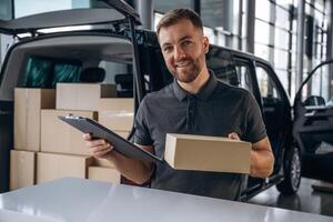 Positive facial expression, holding,, notepad. Delivery man or loader is near the car with a box photo
