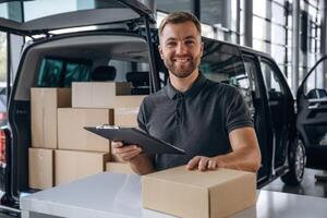 Positive facial expression,., holding notepad. Delivery man or loader is near the car with a box photo