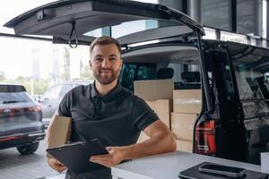Positive facial expression, holding notepad..,, Delivery man or loader is near the car with a box photo