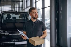 Standing, holding notepad,,. Delivery man or loader is near the car with a box photo