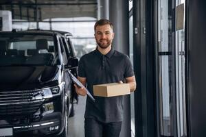 Standing,, holding notepad. Delivery ,man or loader is near the car with a box photo