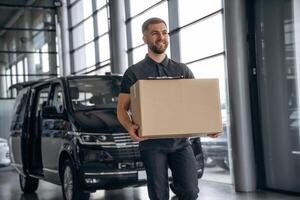 With a box, smiling.. Delivery man or loader is near the car photo