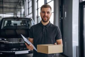 Standing, holding ,,,notepad. Delivery man or loader is near the car with a box photo