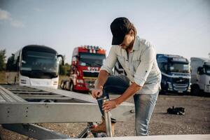 In the casual ,clothes, and in black cap. Truck driver is near the transport outdoors photo