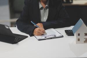 A woman in business attire writing on a notepad with a pen and a house model in the photo