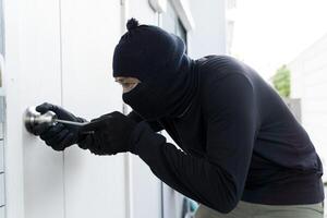 Masked burglar in black clothes using screwdriver to break into house through door lock during daylight robbery photo
