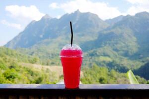Freshly cold Thai red soda water in plastic glass on bamboo fence with beautiful valley Doi Luang Chiangdao mountain view in Chiangmai Province of Thailand. Destination travel photo