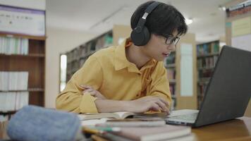 University student in library using laptop and writing notes for online learning video