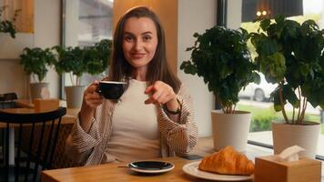 Caucasian woman sitting in cafe female girl drinking tea coffee hey you pointing gesture come here invitation looking at camera sit at table in cafeteria with croissant inviting welcome breakfast photo