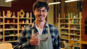 Caucasian man smiling in pottery workshop cheerful happy male potter artisan craftsman guy small business owner sculptor showing thumb up gesture recommend craft store handmade hobby ceramic sculpture photo