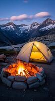 Cozy camping scene with campfire and tent under snowy mountain peaks at dusk photo