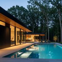 Modern home with pool and deck chairs at dusk, surrounded by forest trees photo