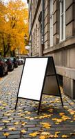 Blank A-Frame Sign on Cobblestone Street with Autumn Leaves and Building Facade photo