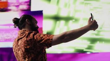 Young carefree man takes selfies and videos surrounded by fun neon lights and beats, representing the fusion of modern DJ culture, media influence and digital art for performance. Camera A.
