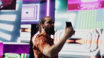 Cool relaxed guy using his smartphone under neon lights, capturing photos and videos as a content creator and DJ. Modern carefree person enjoys visual art in a nightlife scene. Camera A.