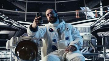 Astronaut taking selfie pictures with smartphone, making hand gestures during planet colonization mission. Orbit station crew member doing photographs with mobile phone during cosmic journey, camera B video