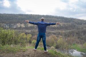 Man stands with arms open wide overlooking a valley on a cloudy day in early spring photo
