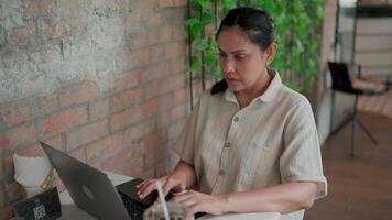 Woman working on laptop in coffee shop. A woman is focused on her work by typing on her laptop with a look of concentration on her face while sitting in a warm coffee shop atmosphere surrounded video